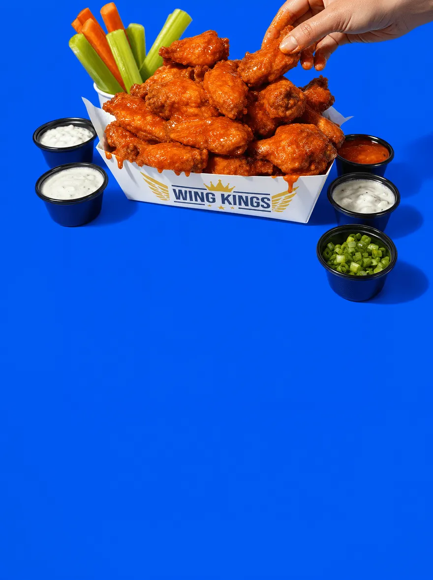 An overhead spread of Wing Kings plates on a blue surface: buffalo wings, dry-rub wings, tenders, chicken sandwiches, crinkle fries, caesar salads, carrots, celery and ranch cups