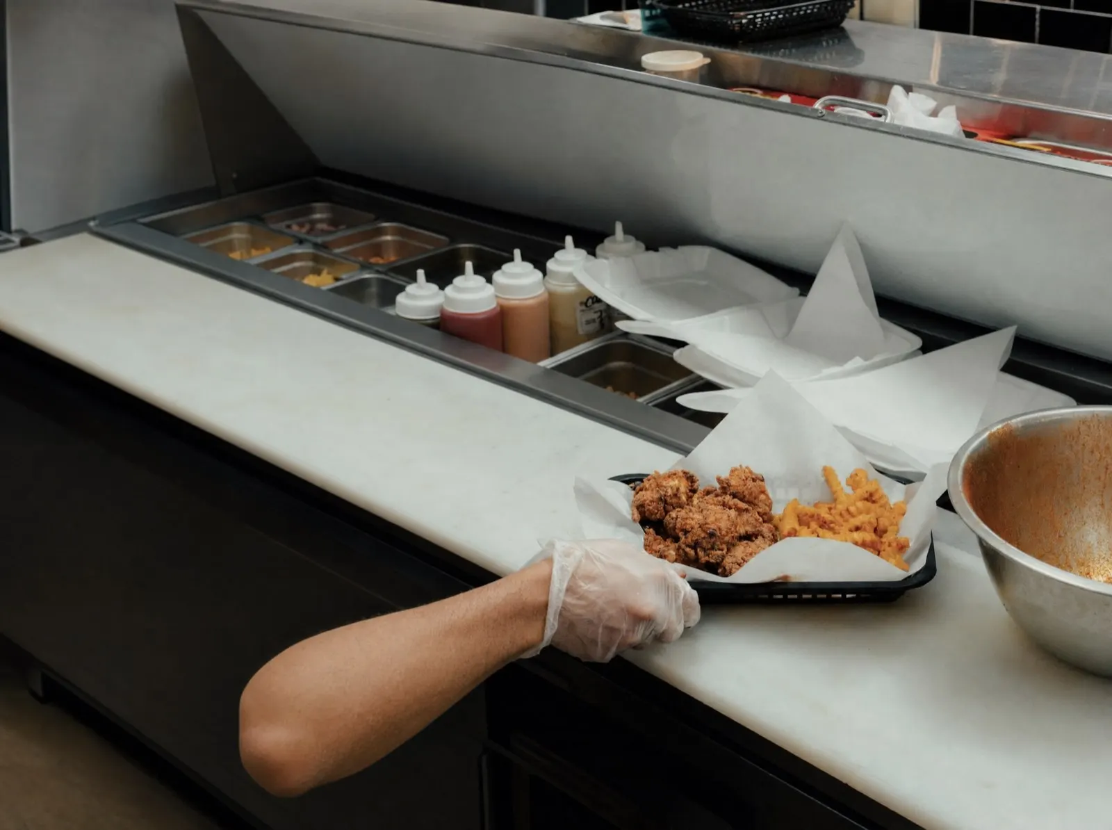 A gloved hand setting a basket of freshly fried tenders and seasoned crinkle fries down on the stainless kitchen line, sauce bottles and prep pans running away behind it