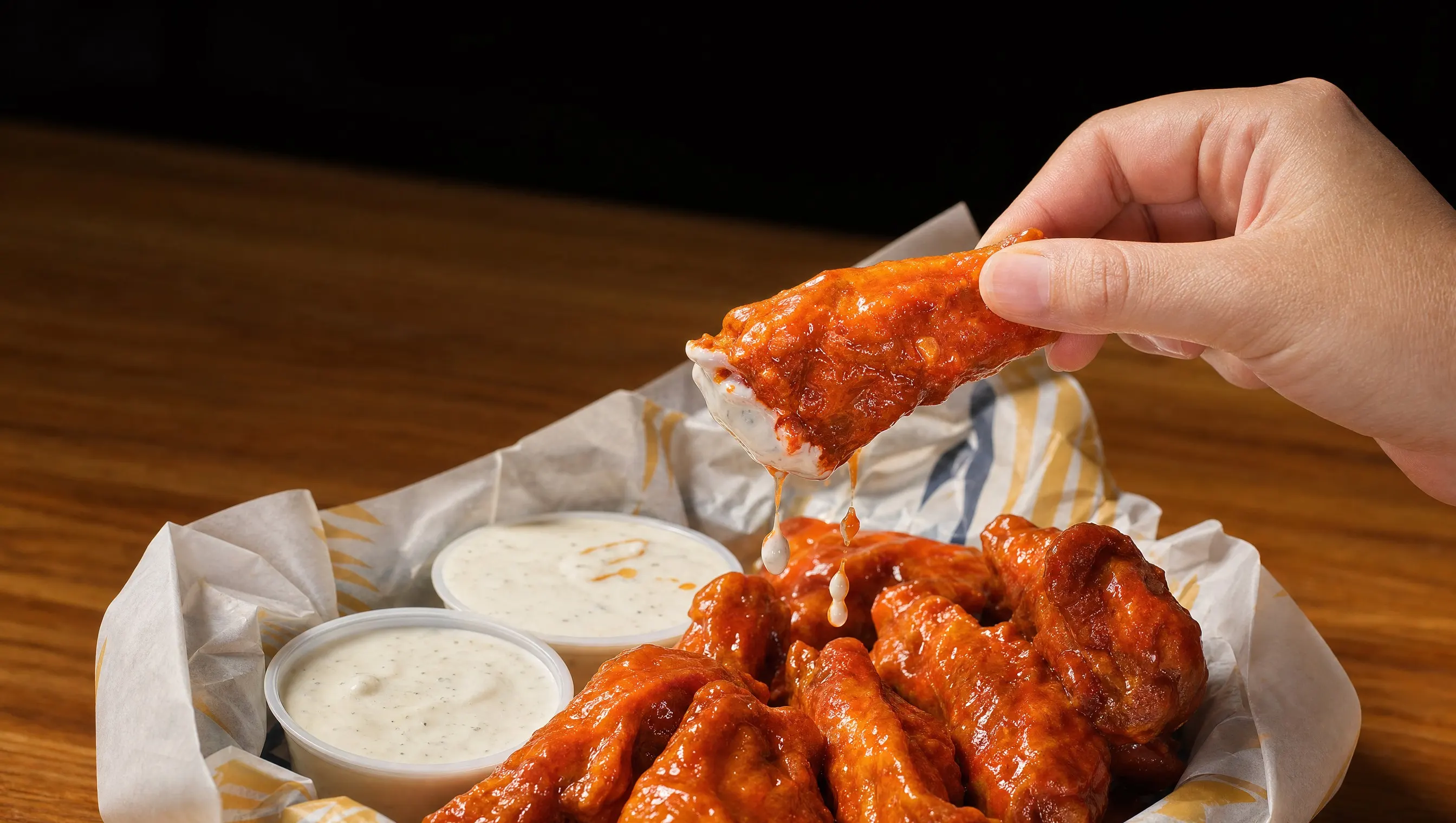 A hand lifting a sauced bone-in wing, its tip dipped in ranch, over a lined basket of wings and two ranch cups