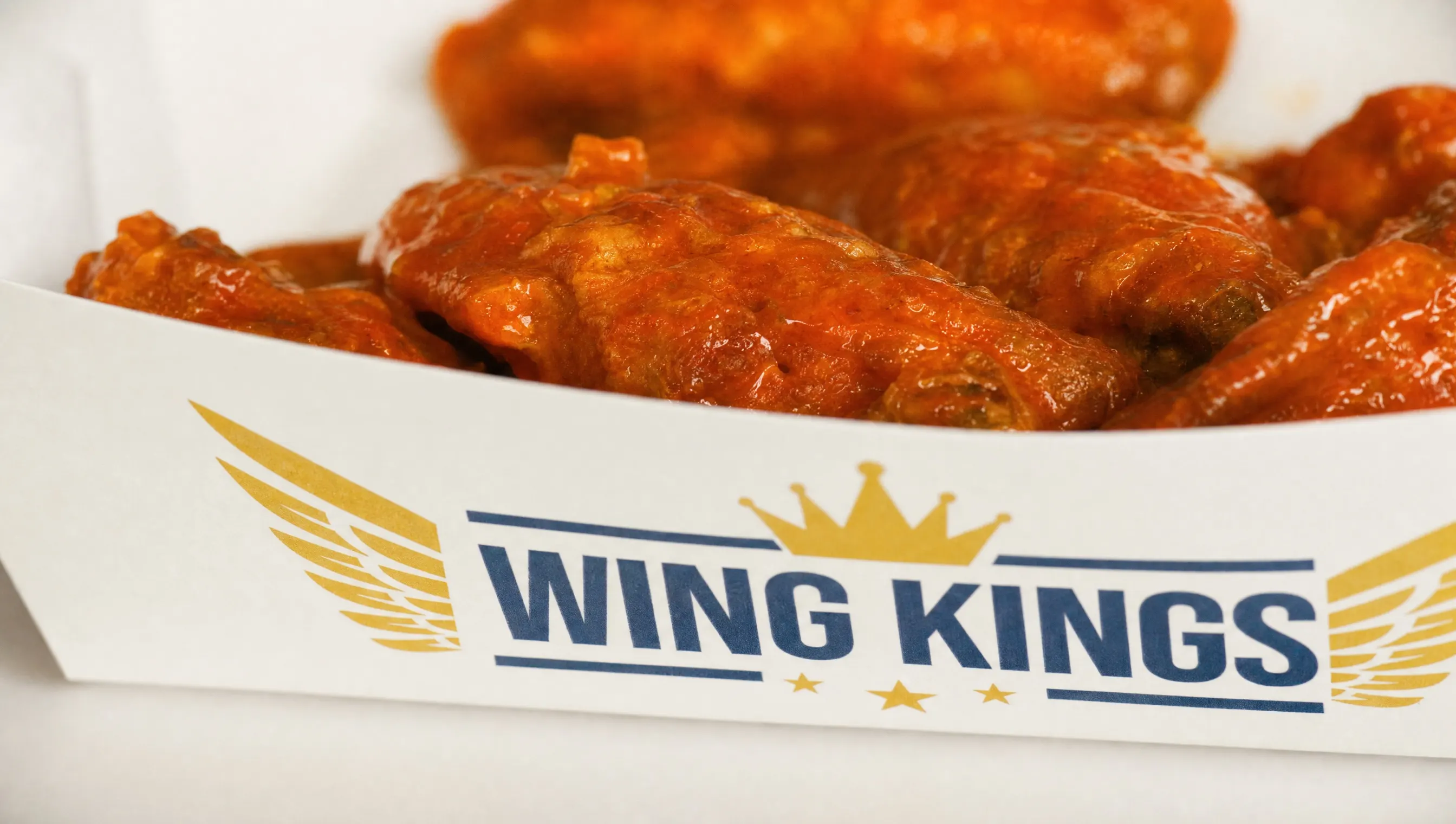 A close crop of bone-in buffalo wings piled above the rim of a branded Wing Kings paper boat, the sauce glossy under hard light