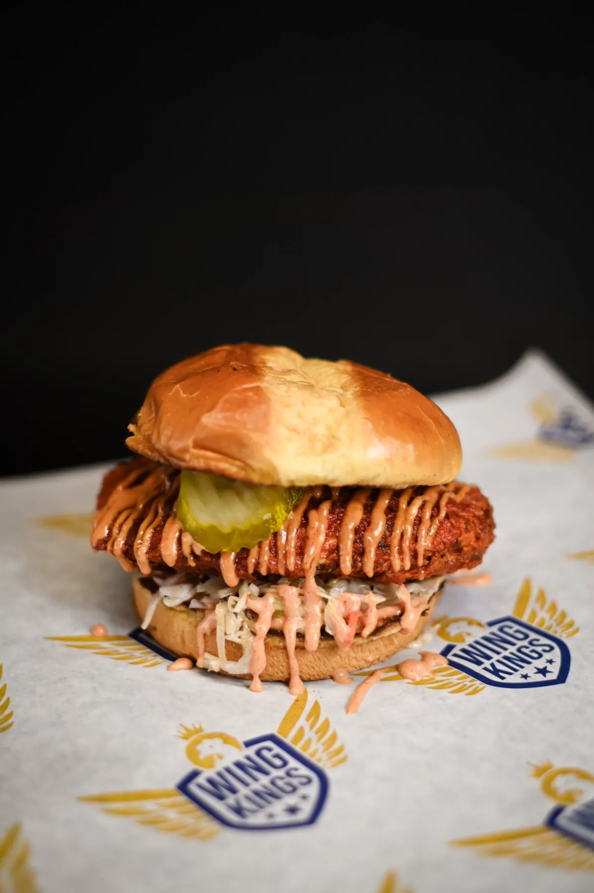 A Nashville chicken sandwich on a buttered potato bun, sauce drizzled over the crispy fillet and running into the slaw, on branded paper against black