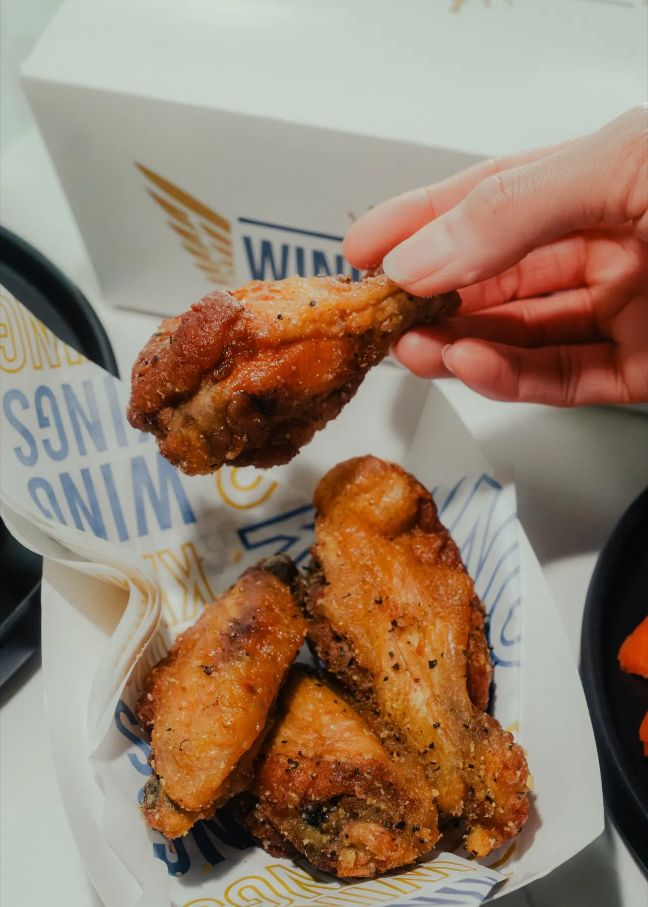 A hand lifting one seasoned bone-in wing out of a branded Wing Kings box, three more stacked in the paper below it
